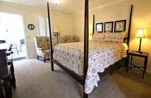 Dark wood four poster bed with red and white patterned bed spread. Nightstands with lamps on either side of bed. Two upholstered chairs off to one side