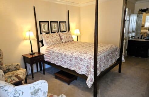 Dark wood four poster bed with red and white patterned bed spread. Nightstands with lamps on either side of bed. Two upholstered chairs off to one side. Bathroom vanity on far side of room.