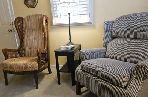 close up of sitting area under window, one high back upholstered cane chair and one plaid armchair
