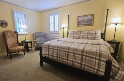 full room view of bed with plaid bedspread, sitting area with two chairs, two windows, two side tables with lamps