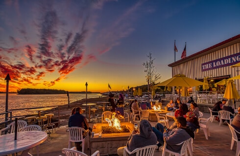 Tables and chairs with people eating, a firepit, a restaurant, all looking over a lake.