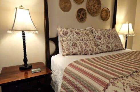 close up of bed and nightstand with lamp, red, sage and cream patterned bed spread, basket decor on wall