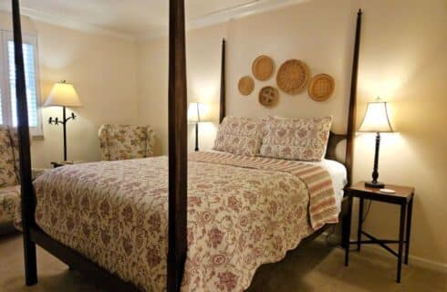 four poster queen bed with red cream and sage patterned bed spread, side tables with lamps on either side, basket decor on wall over bed