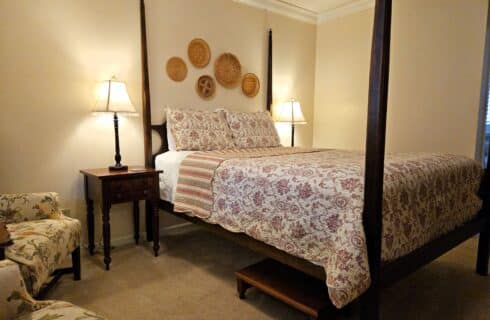 four poster queen bed with red cream and sage patterned bed spread, side tables with lamps on either side, basket decor on wall over bed, step stool under bed