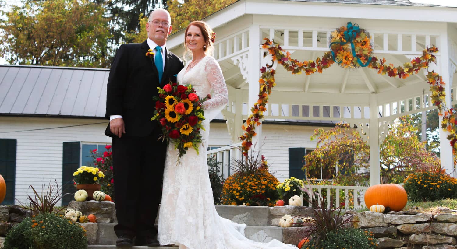 bride holding bouquet with fall flowers and groom standing by white gazebo with draping fall flowers house with green shutters in distance
