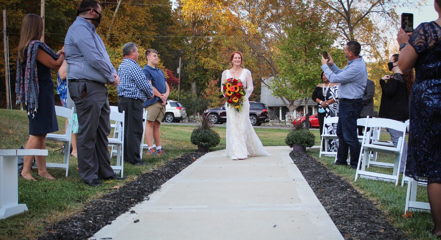 bride walking down aisle with guests standing by white chairs on both sides taking pictures with cell phones