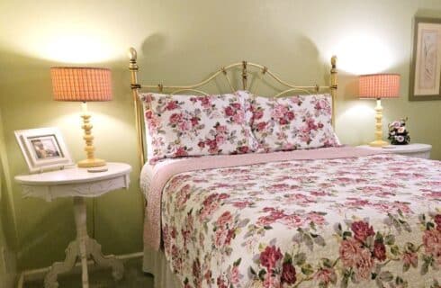 queen bed with brass headboard, white ruffled bed skirt, white and pink floral bed spread, white tables with lamps on either side of bed, light green walls