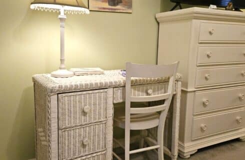 white wicker desk with three drawers, lamp on top, painting of flowers in vase above, white wooden chair, white dresser beside desk, tv on dresser