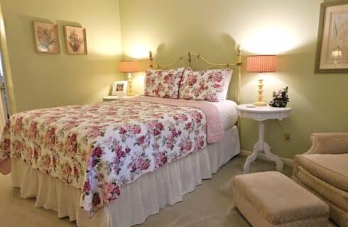 queen bed with brass headboard, white ruffled bed skirt, white and pink floral bed spread, white tables with lamps on either side, bright green walls, framed art, beige armchair with ottoman