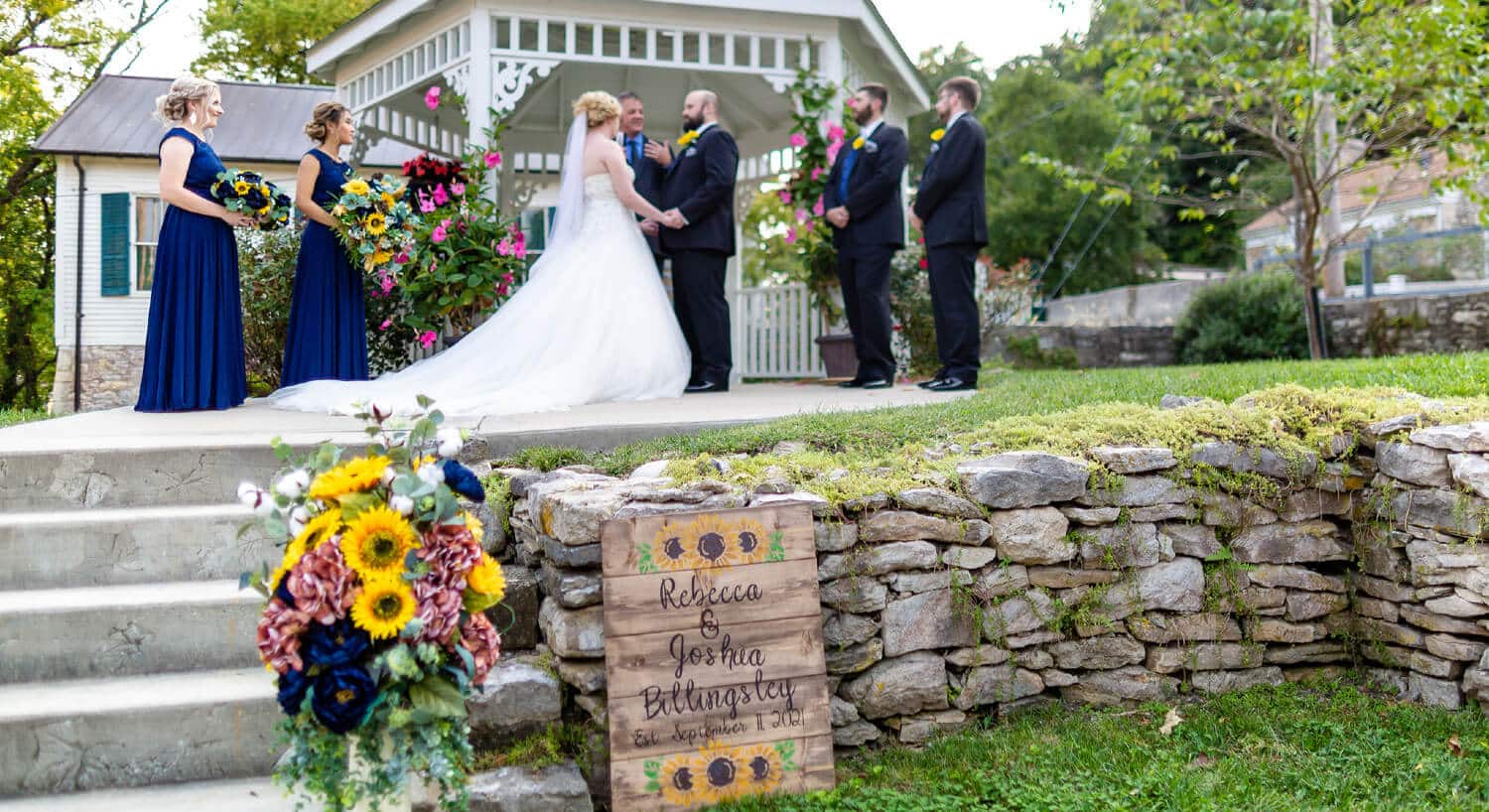Bride and groom holding hands standing in front of minister by gazebo during ceremony with bridal party on each side, wood sign with bride and groom's names