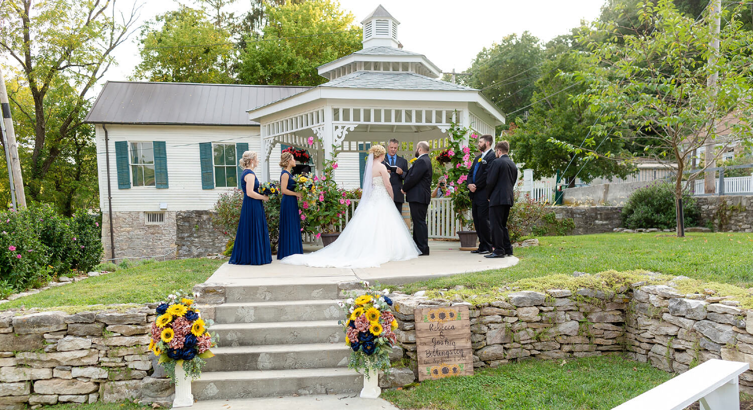 Sunflower arrangements in pots by steps leading up to white gazebo in distance with bride and groom exchanging vows with bridal party on each side.