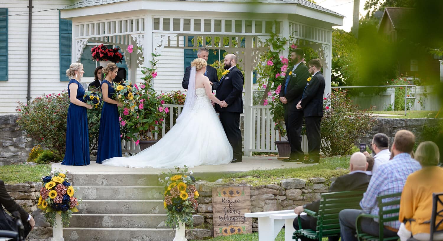 Bridal party standing by white gazebo with minister performing ceremony and guests observing from seating in yard, hanging pink potted flowers.