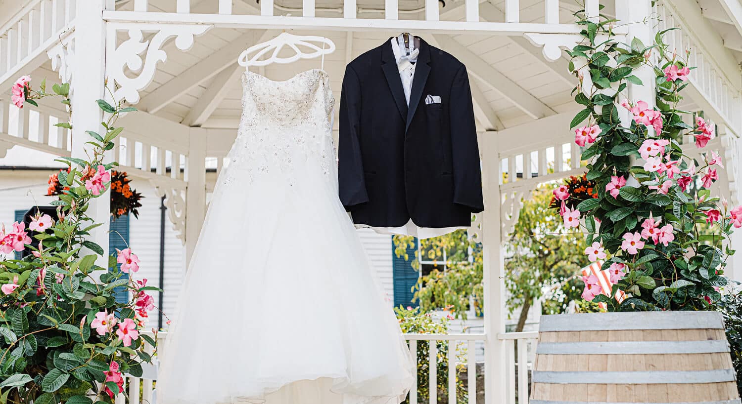 White lacy wedding gown and black tuxedo on hangers hanging from white gazebo. Pink potted flowers on each side of clothing.