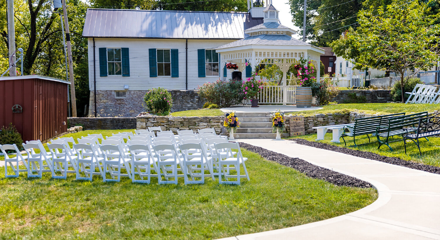 Lawn setup with white folding chairs and benches on each side of cement walk facing white gazebo with hanging potted plants and vases with sunflowers.