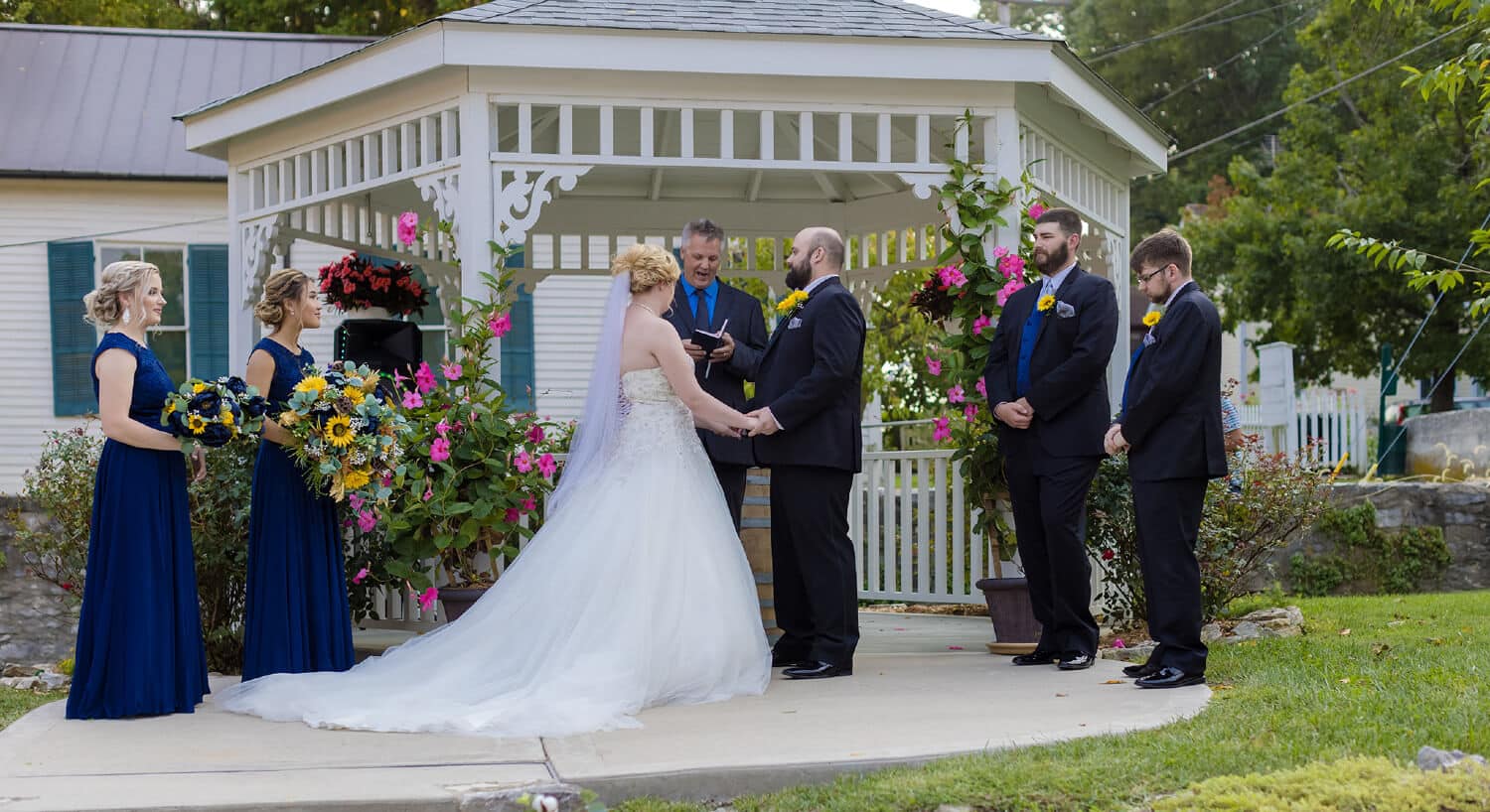 Close-up view of minister performing ceremony with bride and groom holding hands in front of white gazebo with wedding party observing.
