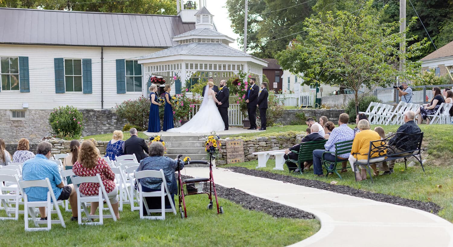 Guests sitting on chairs and benches on each side of cement walkway observing wedding ceremony with bridal party standing in front of white gazebo in distance.