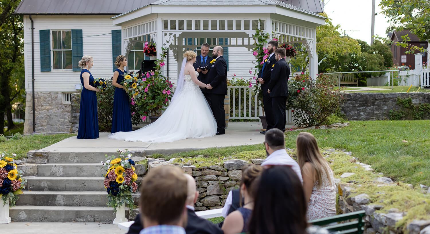 Side view of bride and groom holding hands while exchanging vows by white gazebo with wedding party and guests observing.