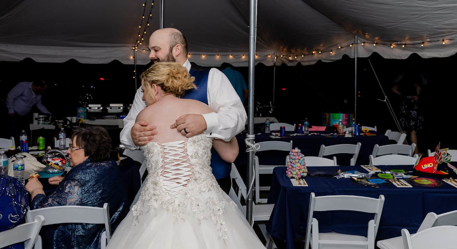 View of bride and grooms hugging while visiting with guests underneath reception tent with tables with blue cloths and white chairs and hanging lights.
