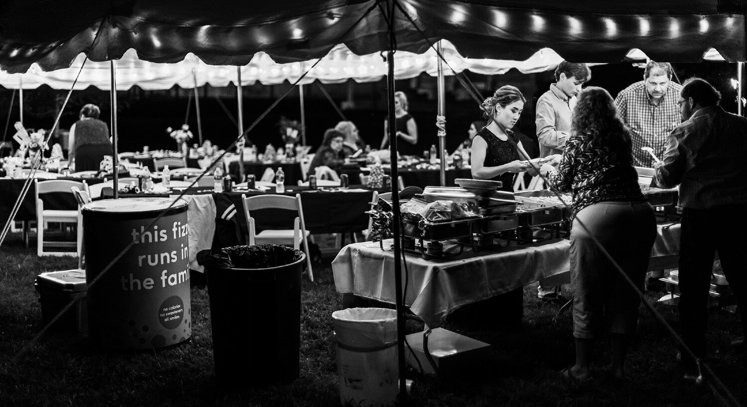 Black and white view of guests congregated underneath tent setup with tables & chairs setup for reception