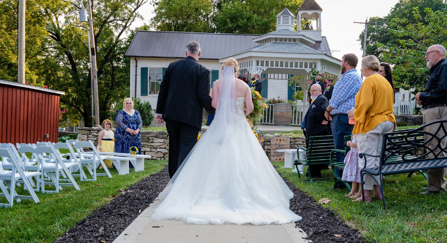 Father walking bride down concrete path toward white gazebo with guests observing from seating area on both sides of walkway.