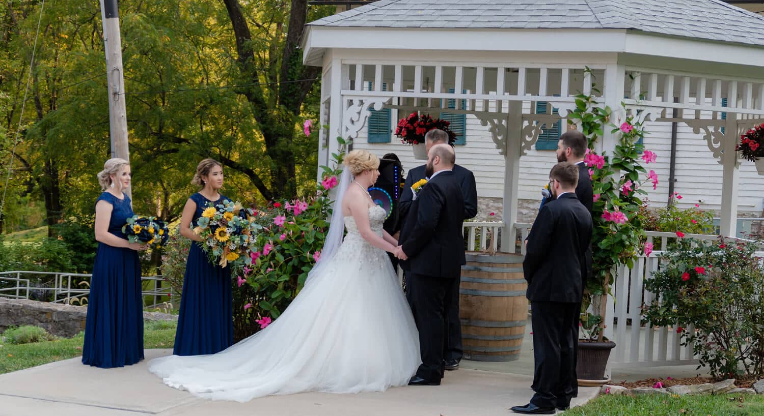 Close-up view of bride and grooms holding hands by gazebo with hanging potted pink flowers during exchanging of vows with wedding party observing.