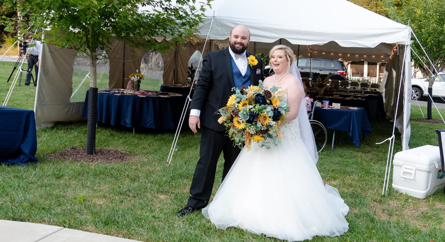 Smiling couple with bride holding large bouquet of sunflowers and others in front of white reception tent setup in yard.