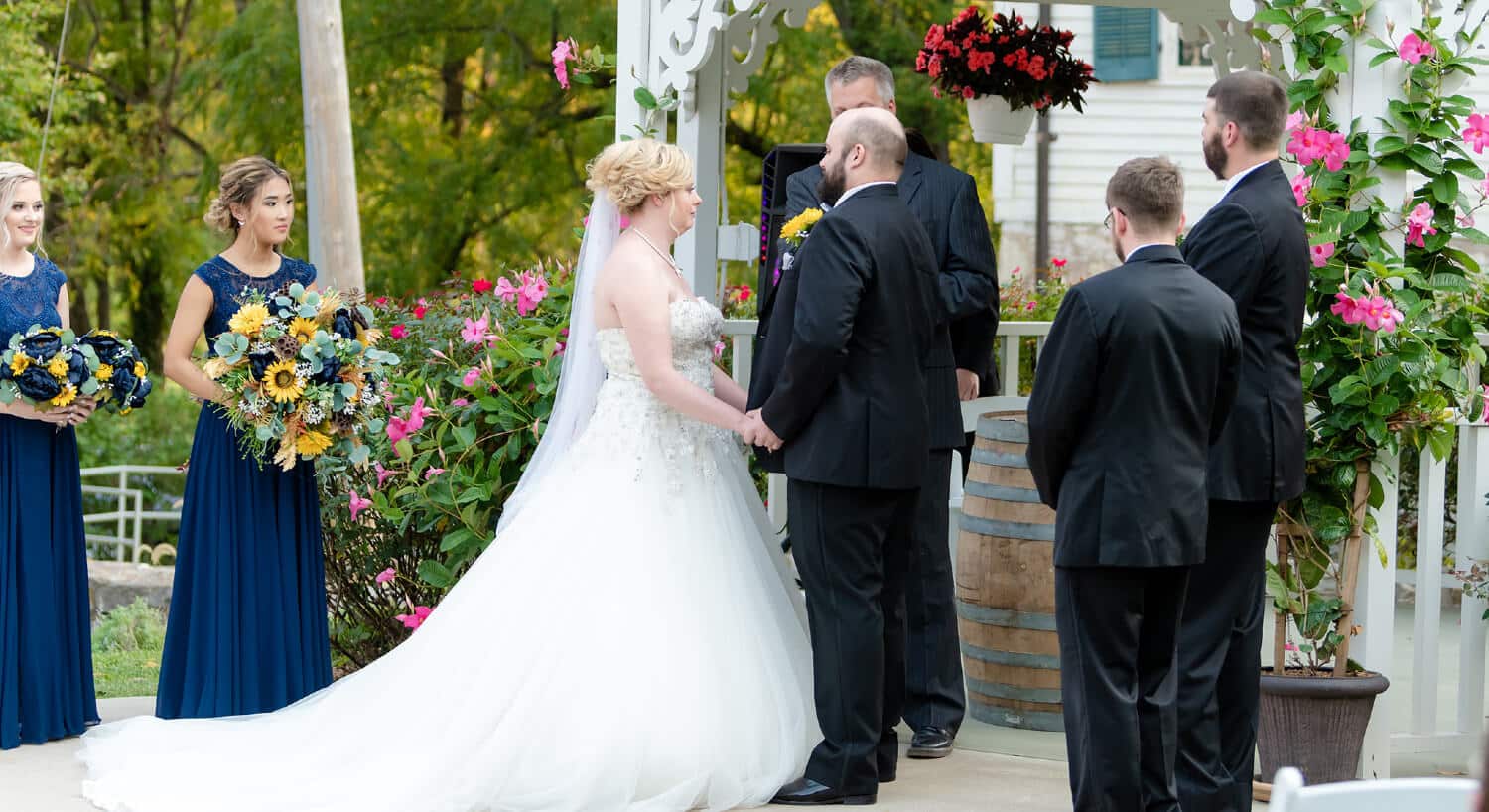 Close-up side view of bride and groom holding hands standing in front of minister with observing wedding party by white gazebo with hanging potted flowers.
