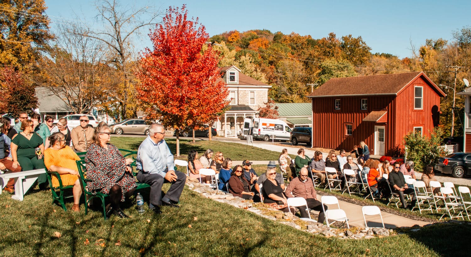 A group of people in chairs and outside preparing for a wedding, with trees with fall foliage of reds, oranges and browns and buildings around the grounds.
